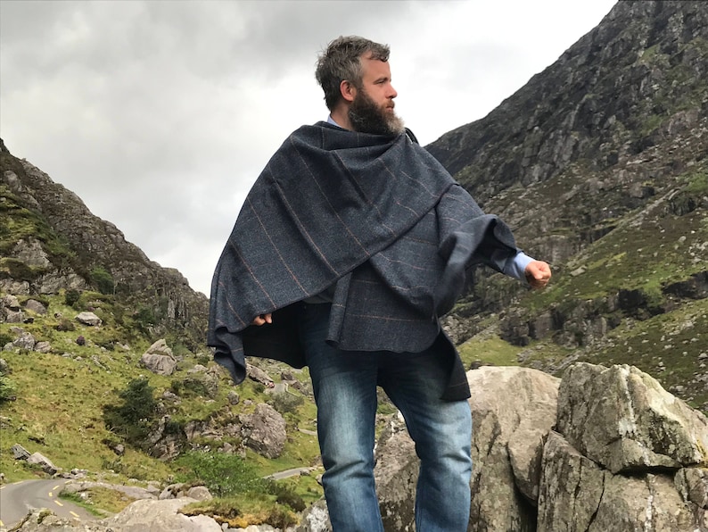 May include: A man wearing a blue and gray plaid poncho standing on a rocky path in a mountainous landscape.