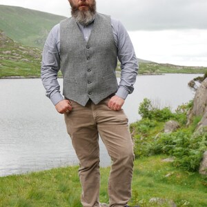 May include: A man wearing a gray plaid vest over a light blue button-down shirt, brown corduroy pants, and black shoes. He is standing on a rock with a lake and mountains in the background.