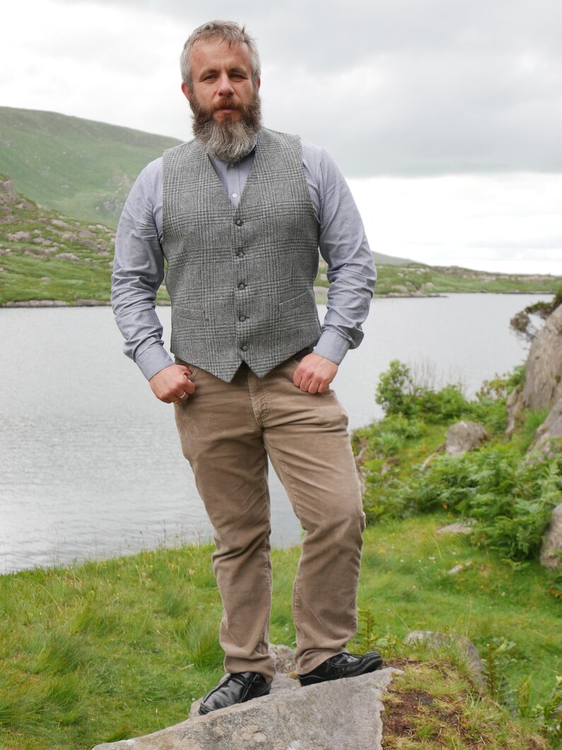 May include: A man wearing a gray plaid vest over a light blue button-down shirt, brown corduroy pants, and black shoes. He is standing on a rock with a lake and mountains in the background.