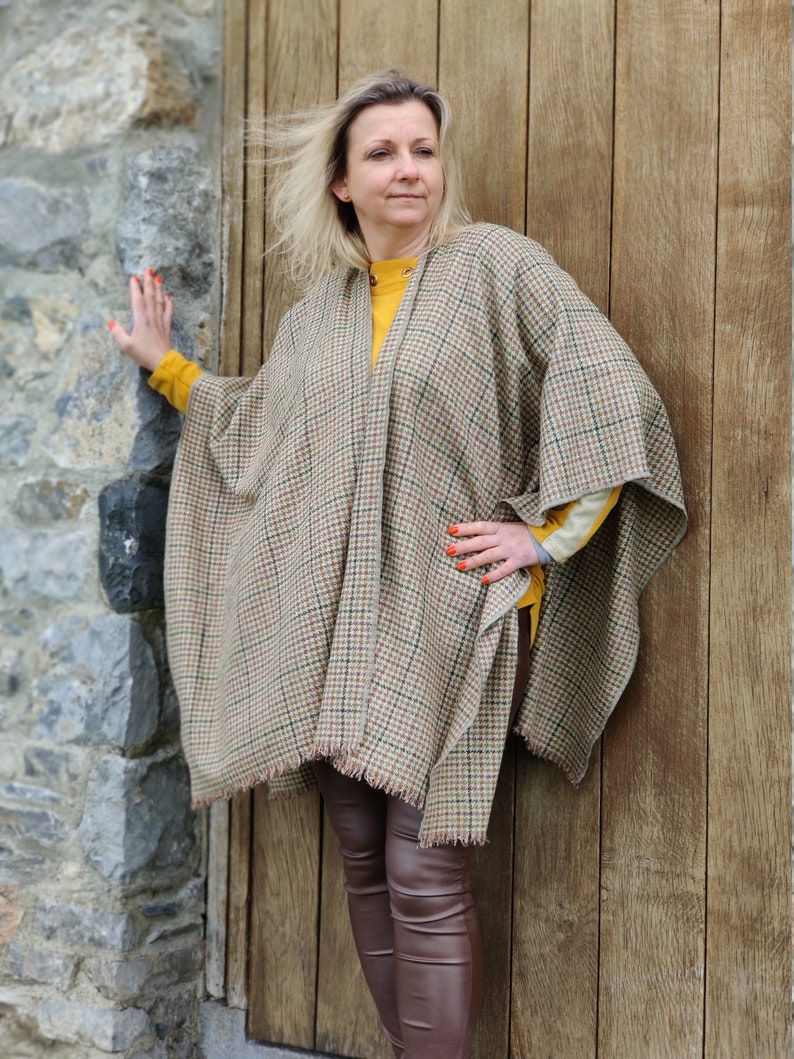 May include: A woman wearing a brown and beige plaid poncho with a fringe trim. She is standing next to a wooden door and has her hand on the door.