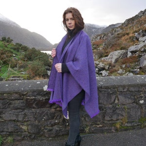 May include: A purple tweed cape with a fringed edge. The cape is worn over a black turtleneck and black pants. The background features a stone wall and a scenic mountain landscape.