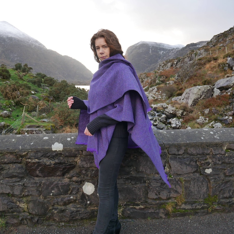 May include: A woman wearing a layered purple cape with a textured weave. The cape has a draped design with a pointed edge. The woman is standing in front of a stone wall with mountains in the background.
