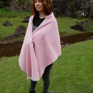 May include: A pink woven wool shawl with a fringe border. The shawl is draped over a person's shoulders in a natural setting.