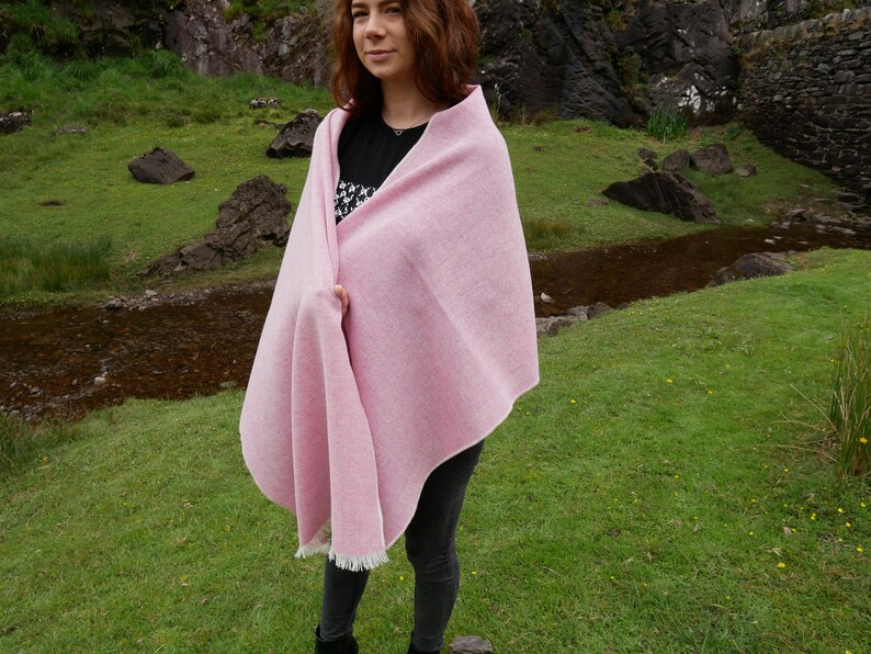 May include: A pink woven wool shawl with a fringe border. The shawl is draped over a person's shoulders in a natural setting.