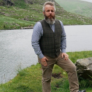 Peut inclure: Un homme barbu portant une chemise boutonn&eacute;e bleue, un gilet &agrave; carreaux brun et vert et un pantalon marron debout devant un lac.