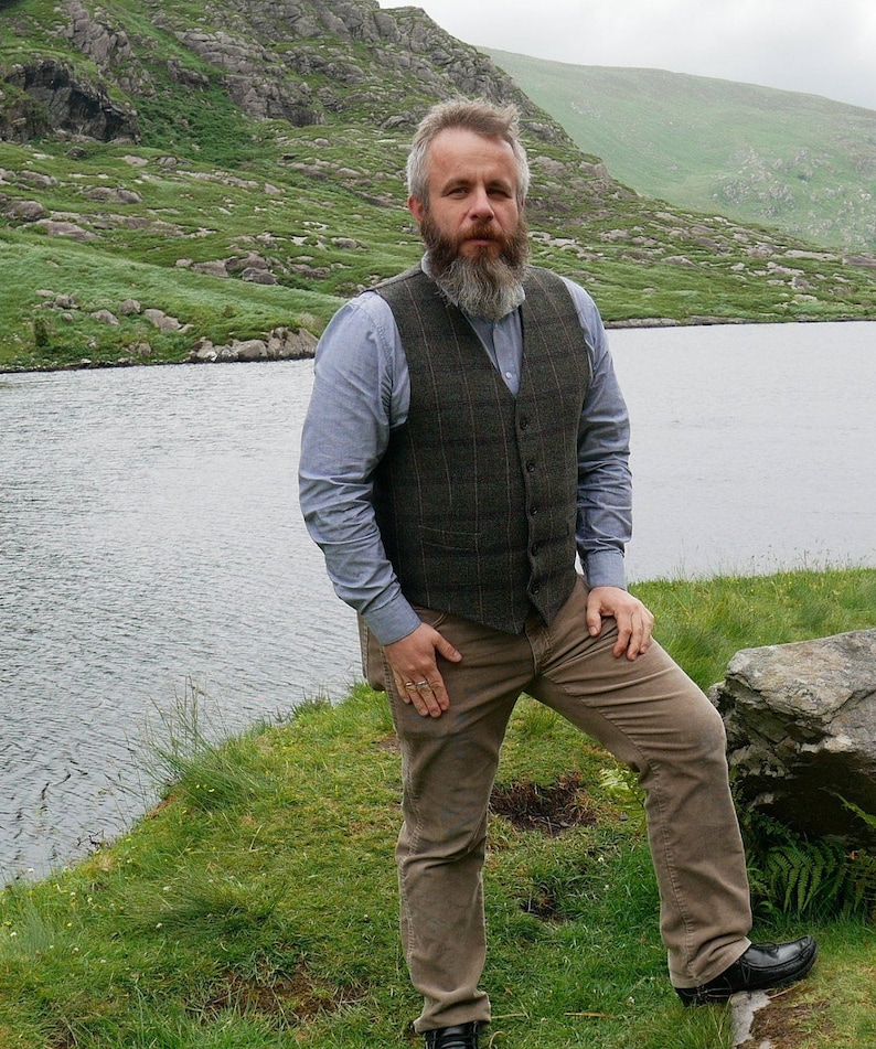 Peut inclure: Un homme barbu portant une chemise boutonn&eacute;e bleue, un gilet &agrave; carreaux brun et vert et un pantalon marron debout devant un lac.