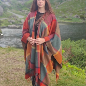 May include: A woman wearing a plaid poncho with fringe. The poncho is in shades of brown, orange, red, and blue. The woman is standing in a grassy field with a lake and mountains in the background.