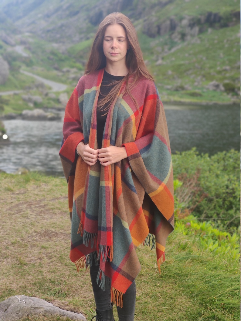 May include: A woman wearing a plaid poncho with fringe. The poncho is in shades of brown, orange, red, and blue. The woman is standing in a grassy field with a lake and mountains in the background.