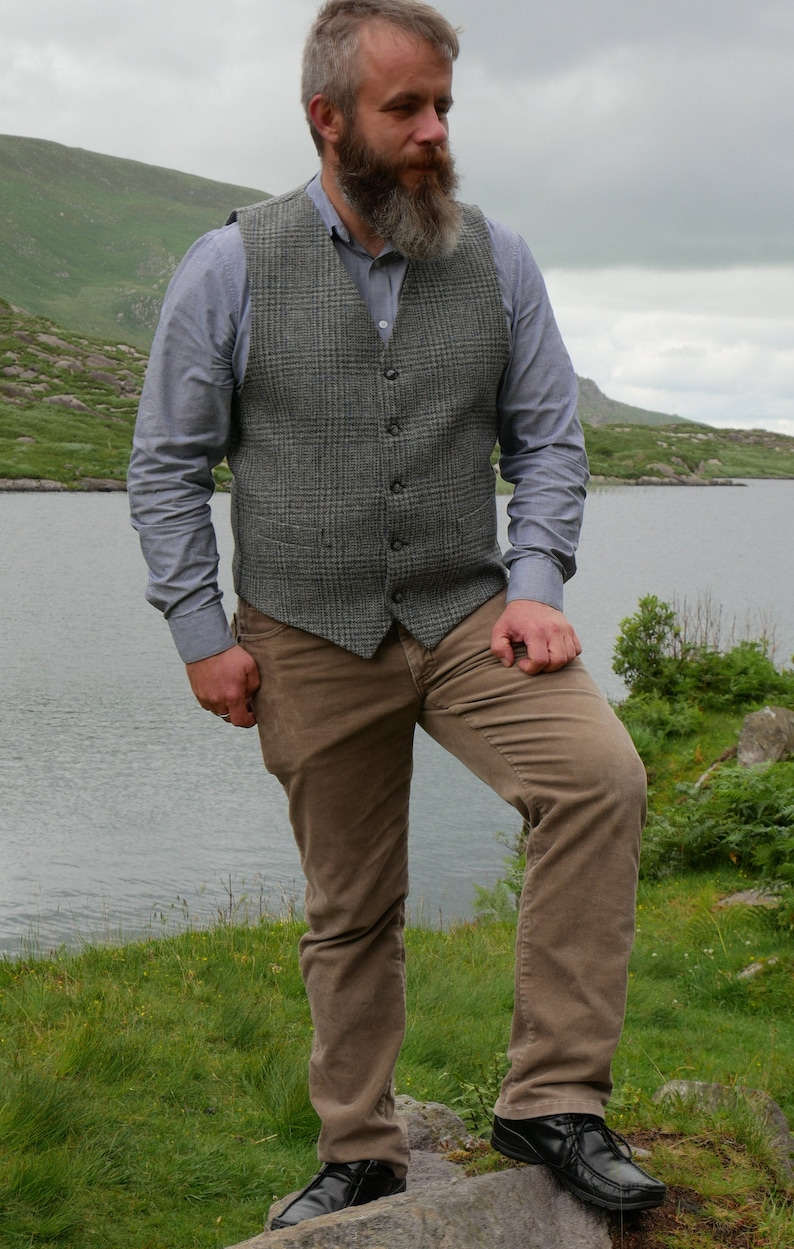 May include: A man wearing a gray plaid vest over a light blue button-down shirt and brown corduroy pants. He is standing on a rock by a lake.