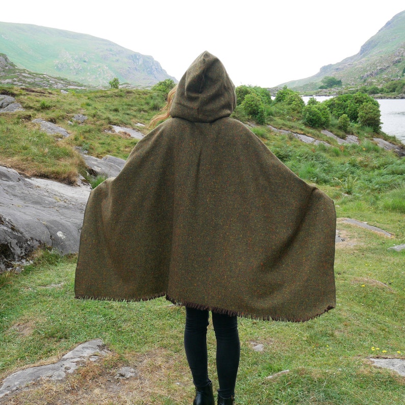May include: A green hooded cloak with a fringed edge. The cloak is made of a thick, textured fabric and is worn by a person standing in a grassy field.