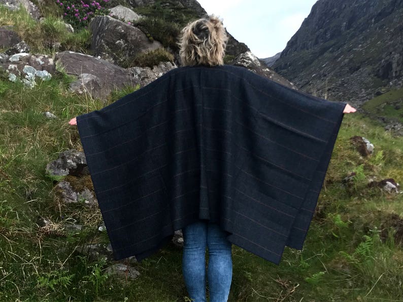 May include: A dark blue plaid poncho with a fringed edge. The poncho is worn by a person standing in a mountainous landscape.
