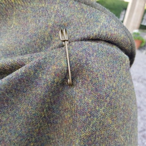 May include: A close-up of a green, brown, and yellow tweed fabric with a gold safety pin.