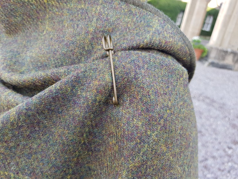 May include: A close-up of a green, brown, and yellow tweed fabric with a gold safety pin.
