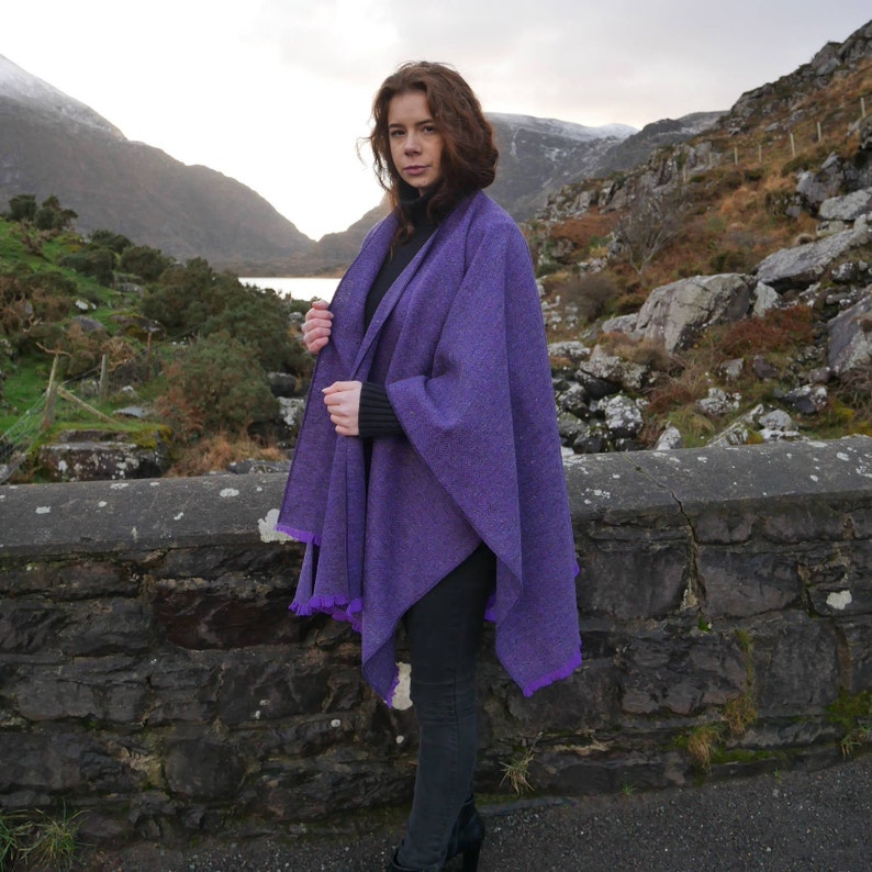 May include: A purple cape with a fringed edge. The cape is made of a tweed-like material and is worn over a black turtleneck. The background features a stone wall, mountains, and a lake.