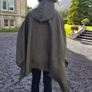 May include: A hooded green wool poncho with a frayed edge. The poncho is worn by a person standing in front of a stone building.