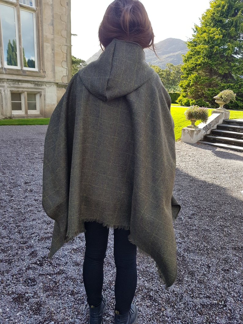 May include: A hooded green wool poncho with a frayed edge. The poncho is worn by a person standing in front of a stone building.