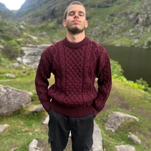 May include: A burgundy crew neck sweater with a textured cable knit pattern. The sweater is worn with black cargo pants. The background features a lake and mountains.