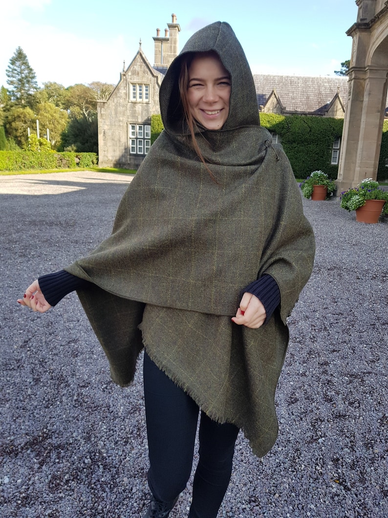 May include: A green and brown hooded poncho with a fringed edge. The poncho is made of a tweed fabric and has a relaxed fit.