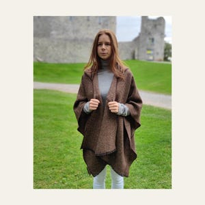 May include: A brown and black herringbone patterned hooded poncho worn over a light gray long-sleeved turtleneck shirt. The poncho features an open front design with a textured weave and dark edges, providing a layered look.