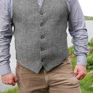 May include: A gray tweed vest with a button-up closure. The vest has a plaid pattern and is worn over a light blue button-down shirt. The person is wearing brown corduroy pants.