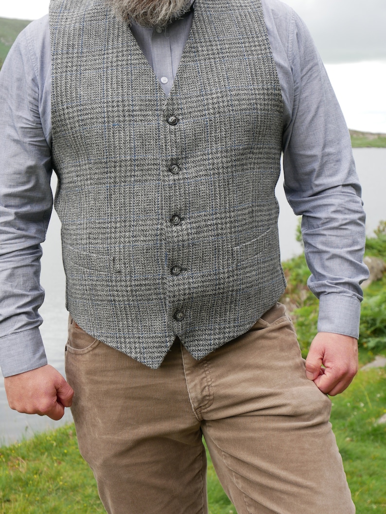 May include: A gray tweed vest with a button-up closure. The vest has a plaid pattern and is worn over a light blue button-down shirt. The person is wearing brown corduroy pants.