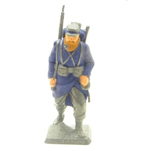 May include: A blue painted toy soldier figurine of a French infantryman wearing a blue uniform, a gray hat, and a backpack. The soldier is holding a rifle and has a pipe in his mouth. The figurine is standing on a base with the words "Soldat de la Marine" written on it.
