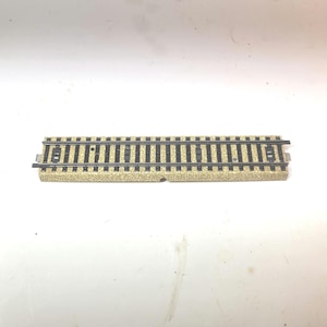 May include: A straight model train track section featuring two parallel dark gray metal rails mounted on a textured, light tan plastic base that simulates gravel ballast, with black plastic ties spaced evenly along the entire length.