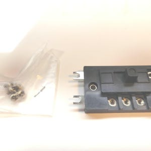 May include: A black electrical component with silver metal prongs and screw terminals. A small plastic bag contains several small, metallic screws. The component has a rectangular shape with a raised section and several holes.