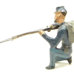 May include: A vintage toy soldier in a kneeling position, aiming a rifle. The figure is painted in shades of blue, gray, and tan, with a backpack and a hat. The toy soldier is a collectible item.