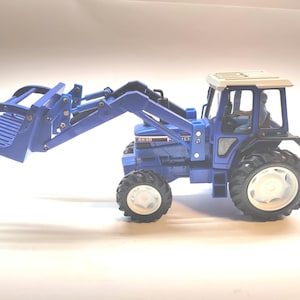Puede incluir: Un tractor de juguete azul con un cargador frontal y neumáticos grandes. El tractor tiene un techo blanco y una cabina detallada. El cargador frontal está levantado, y el tractor está sobre un fondo blanco.