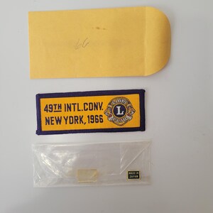 May include: A rectangular embroidered patch with a yellow center and dark blue border. The text reads 49TH INTL.CONV. NEW YORK, 1966 alongside a Lions Club logo. Included are a yellow paper envelope with the number 66 and a small Made in Japan pin.