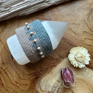Peut inclure: Un cristal de sélénite clair et pointu est orné d'un macramé bleu et beige, orné de perles en bois. Le cristal repose sur une surface en bois, accompagné de fleurs séchées, créant une esthétique naturelle et terreuse.