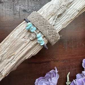 May include: Two handmade bracelets. One is a wide, woven macrame style in a neutral beige colour. The other is a beaded bracelet with turquoise-coloured stones and a silver-toned charm. The bracelets are displayed on a piece of driftwood.