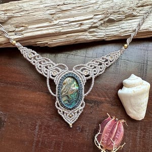 May include: A handmade macramé necklace with an oval abalone shell pendant. The necklace has a grey cord with detailed knotting and gold-coloured beads. A seashell and dried seed pod are also visible.