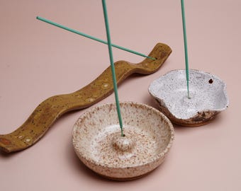 May include: A selection of ceramic incense holders in different designs. The set features a long, curved holder and two speckled bowls, in shades of brown and white. Teal incense sticks are placed in the holders.