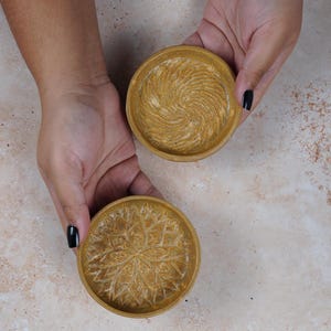 May include: Two small, round, golden-brown ceramic dishes with decorative patterns. One dish has a swirling pattern, and the other has a floral pattern.