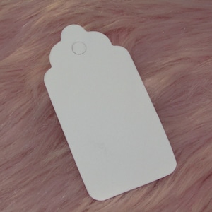 May include: A blank white gift tag with a scalloped edge and a hole punched in the top. The tag is lying on a pink fluffy surface.