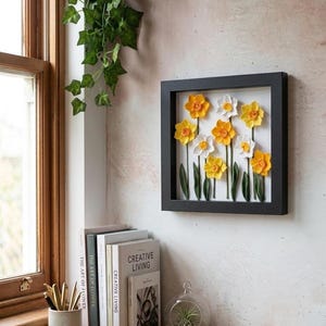Peut inclure: Un cadre carré noir de type boîte vitrine contenant une composition florale en relief de jonquilles jaunes et blanches avec des tiges et des feuilles vertes sur un fond blanc. L'œuvre est fixée au mur à côté d'un cadre de fenêtre en bois.