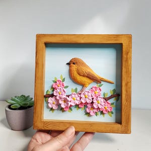 Op de afbeelding: Een vierkante houten lijst met een driedimensionaal reliëf van een oranje vogel die op een bruine tak met roze bloemen zit. De achtergrond is een lichtblauwe lucht. De vogel heeft een donkere snavel en gestructureerde veren, de bloemen zijn roze.