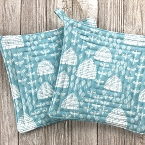 Puede incluir: Dos agarraderas de olla acolchadas azules con un patrón de colmena y libélula blanco.