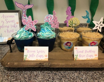 May include: A wooden tray with two types of desserts. One is blue jello cups with a mermaid tail and starfish topper, labeled "Ocean Water Jello Cups". The other is a sand pudding dessert with an oreo crust, topped with a mermaid and a starfish, labeled "Sand Pudding Oreo Cups".