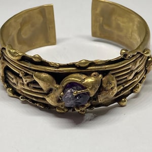 May include: A gold-toned cuff bracelet with an open design. The bracelet features intricate relief detailing and a central, irregular-shaped purple gemstone. The metal has a slightly aged appearance, suggesting a vintage or antique style.