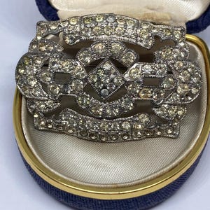 Könnte beinhalten: Vintage silberfarbene Brosche mit klaren Strasssteinen in einer blauen Schmuckschatulle. Die Brosche hat ein dekoratives Design mit einer rautenförmigen Mitte und einem Banner oben. Die Schatulle hat einen goldenen Rand.