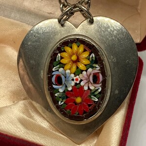 May include: A silver-toned heart-shaped pendant with a mosaic floral design. The mosaic features yellow, red, blue, and pink flowers. The pendant hangs from a silver chain and is displayed in a red velvet box.