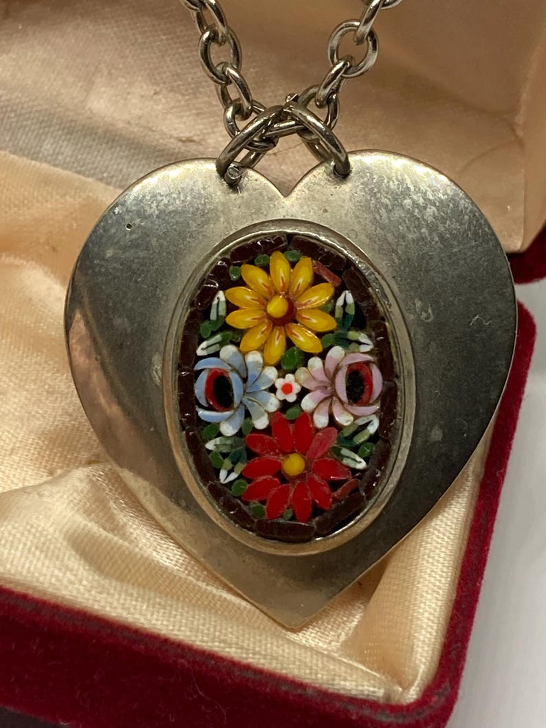 May include: A silver-toned heart-shaped pendant with a mosaic floral design. The mosaic features yellow, red, blue, and pink flowers. The pendant hangs from a silver chain and is displayed in a red velvet box.