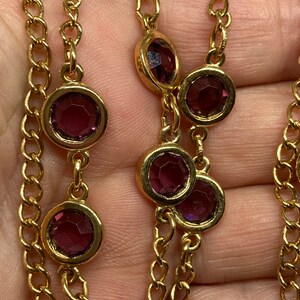 May include: A gold-toned necklace featuring a chain design with alternating round, bezel-set, purple gemstones. The gemstones are evenly spaced along the chain, creating a delicate and elegant look. The necklace is shown against a neutral background.