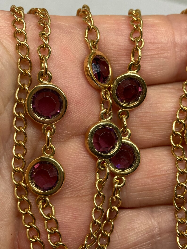 May include: A gold-toned necklace featuring a chain design with alternating round, bezel-set, purple gemstones. The gemstones are evenly spaced along the chain, creating a delicate and elegant look. The necklace is shown against a neutral background.