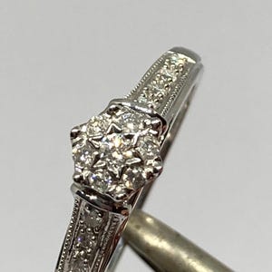 May include: A close-up of a silver ring with a cluster of small, sparkling diamonds at the center. The band features smaller diamonds and detailed engravings. The ring is set against a plain background, highlighting its intricate design.