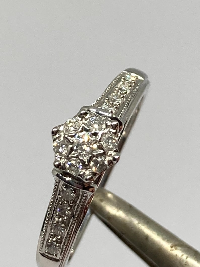 May include: A close-up of a silver ring with a cluster of small, sparkling diamonds at the center. The band features smaller diamonds and detailed engravings. The ring is set against a plain background, highlighting its intricate design.