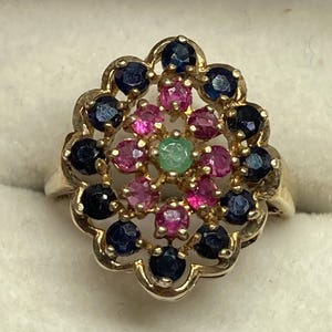 May include: A gold ring featuring a central green gemstone surrounded by a cluster of pink gemstones, with an outer scalloped border set with dark blue gemstones. The design creates a floral pattern with multiple round-cut stones in a metal setting.